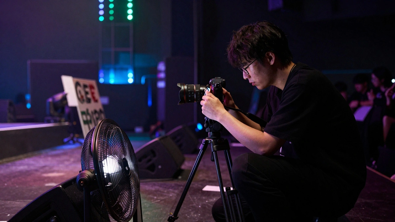 A photographer in black quietly swapping memory cards between songs, stage lights casting shadows on equipment and a fan holding a sign in the foreground.
